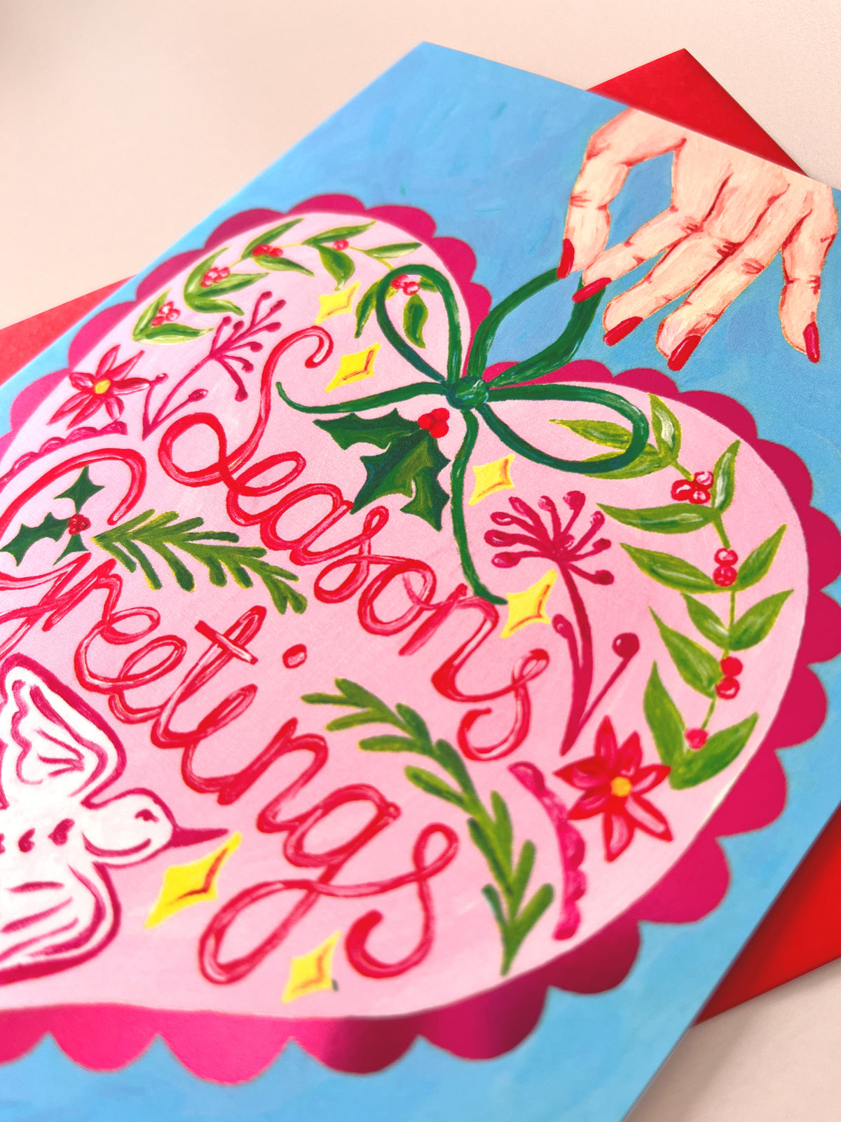 A close-up of a colorful hand-painted Christmas greeting card featuring the words 'SeasonsGreetings' in red cursive script inside a pink heart, decorated with green holly leaves, red berries, and yellow stars, layered over a bright blue background with a hand illustration at the top right.