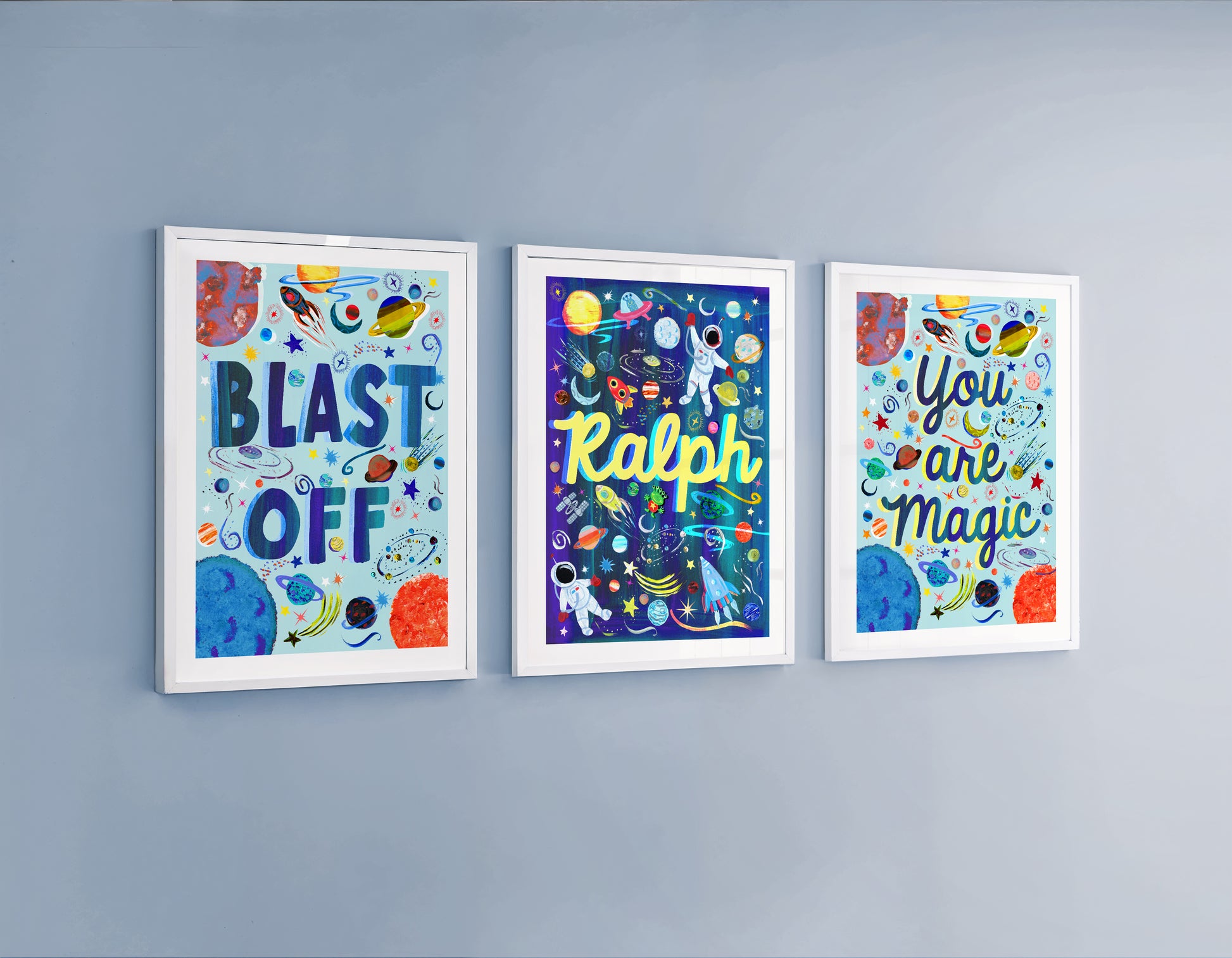 A set f 3 artworks on a blue wall one say 'Blast Off' a personalised name print and the last print displaying words 'you are magic' all designed in a space theme with hand painted space elements of planets, rockets and astronauts.