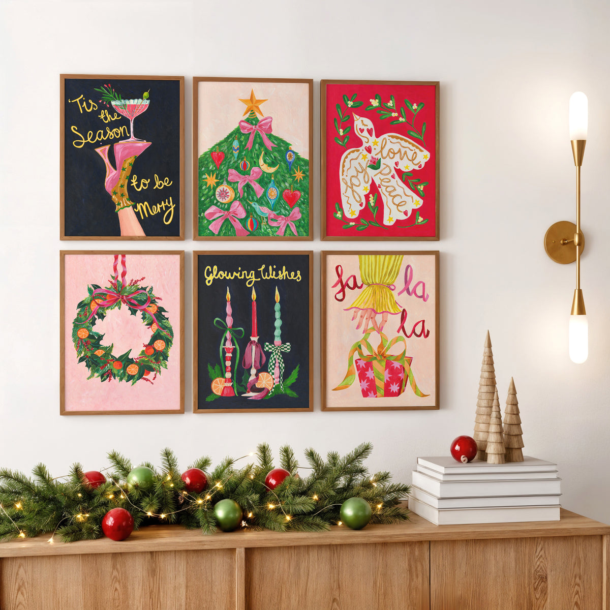 Six framed Christmas art prints arranged in a 2x3 grid on a white wall above a wooden console table. The prints feature festive illustrations and phrases including 'Tis the Season to be Merry,' a decorated Christmas tree, a white bird on red, a green wreath, glowing candles, and 'la la la.