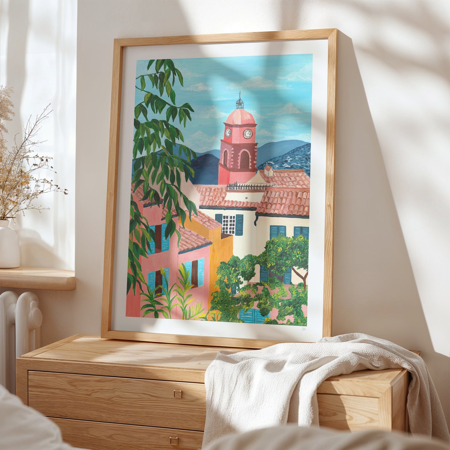 Framed artwork of a colorful village scene on a wall above a wooden dresser.