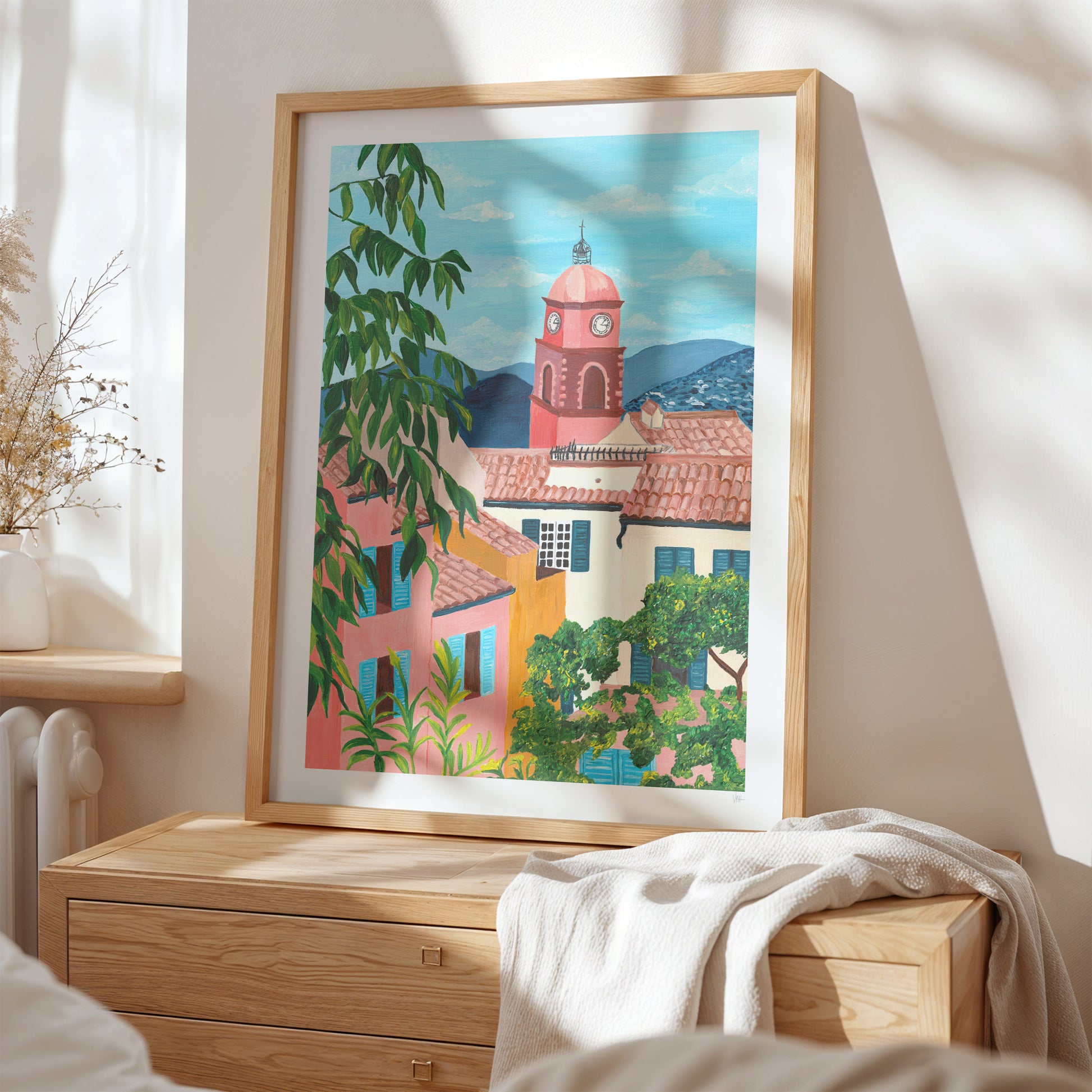 Framed artwork of a colorful village scene on a wall above a wooden dresser.
