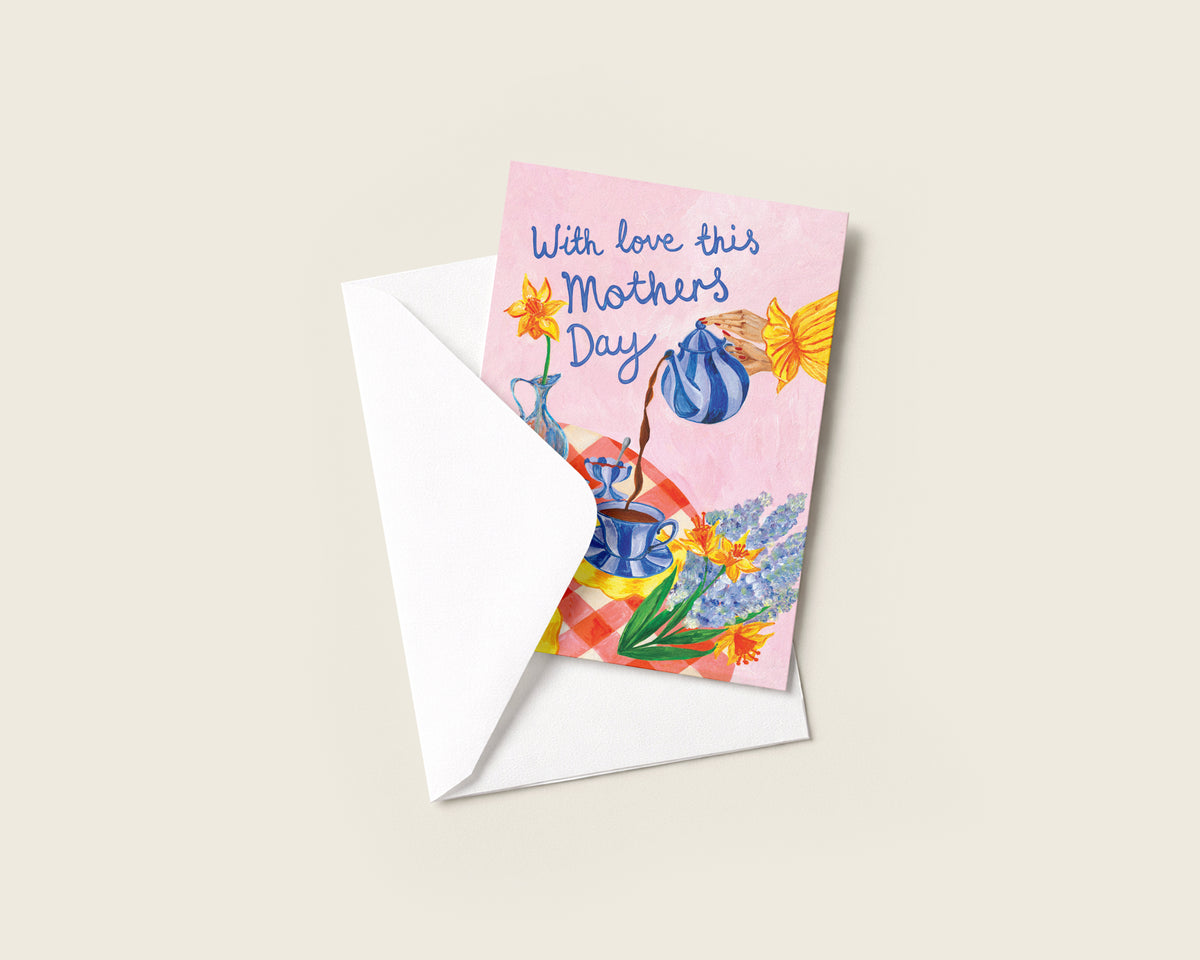 A square Mother's Day greeting card with a pink watercolor background and colorful floral illustrations, partially tucked into a white envelope on a light beige background.