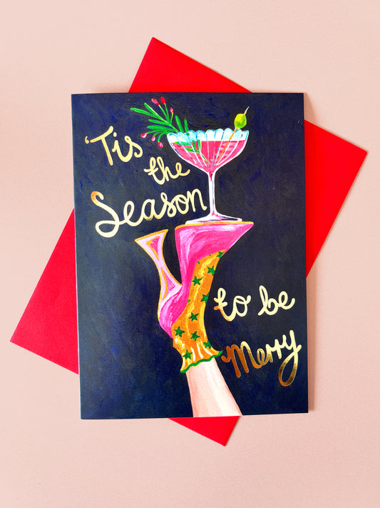 A square greeting card with a dark navy blue background, featuring a hand holding a pink martini glass and the text 'Tis the Season to be Merry' in white and gold lettering. The card is styled on top of red envelopes against a solid pale pink background.