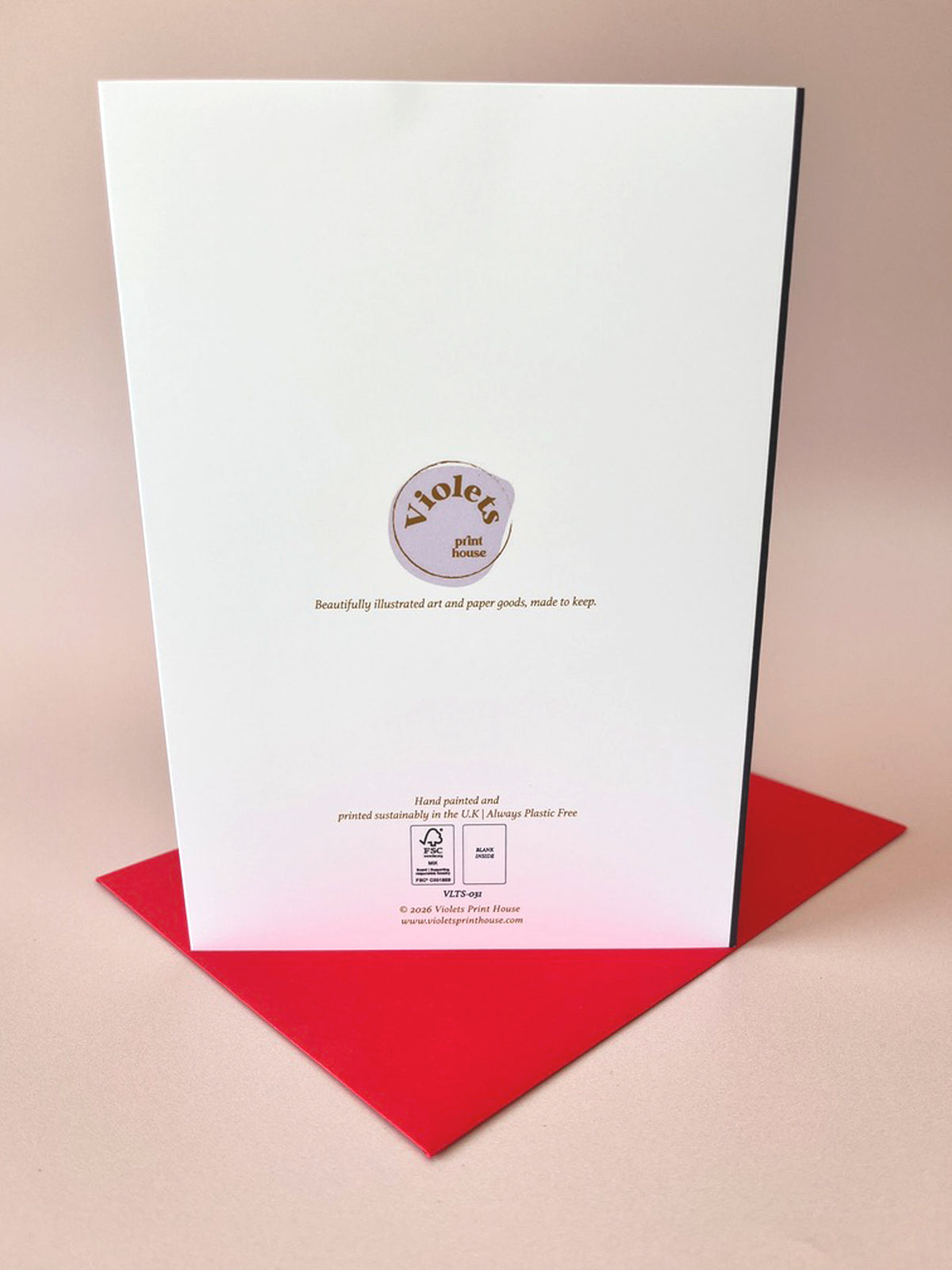 A white card or booklet standing upright on a red surface against a warm beige background. The front has a circular logo reading 'Violets piano music' and small printed text near the bottom.