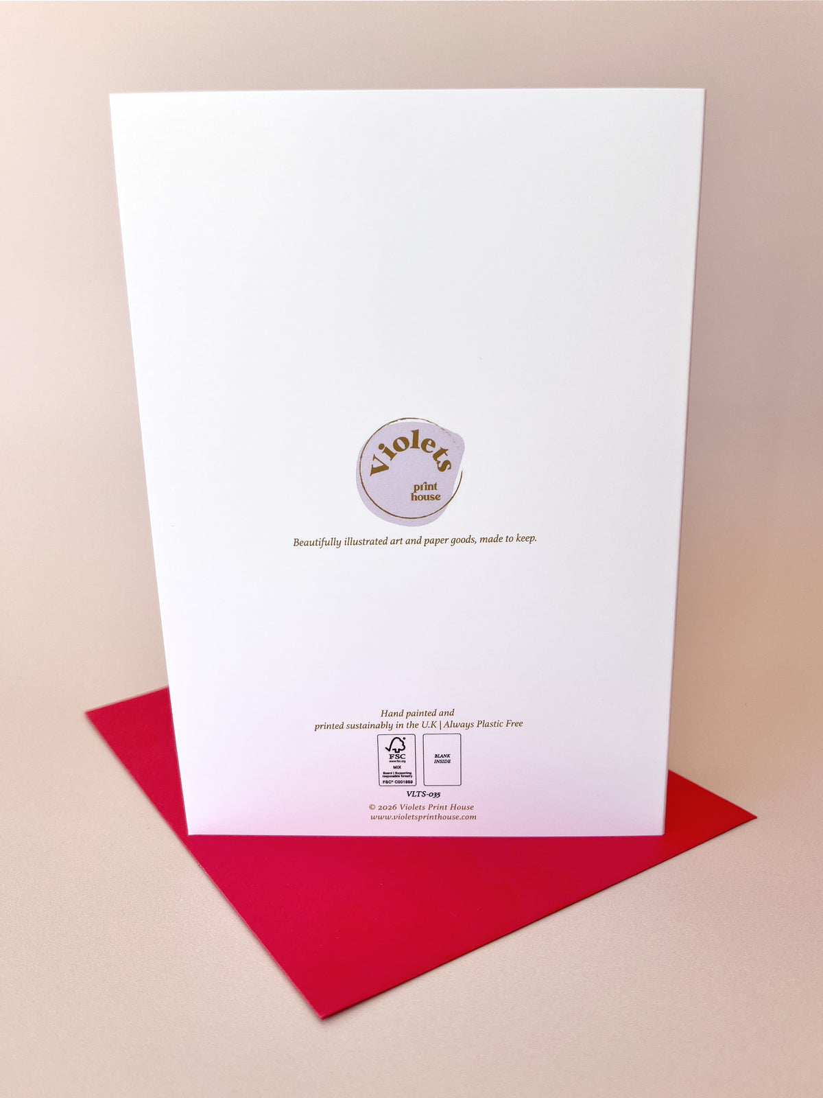 A white art print card with a circular logo for 'Violets printed paper' and a small plastic bag graphic, standing upright on a red envelope against a warm beige background.