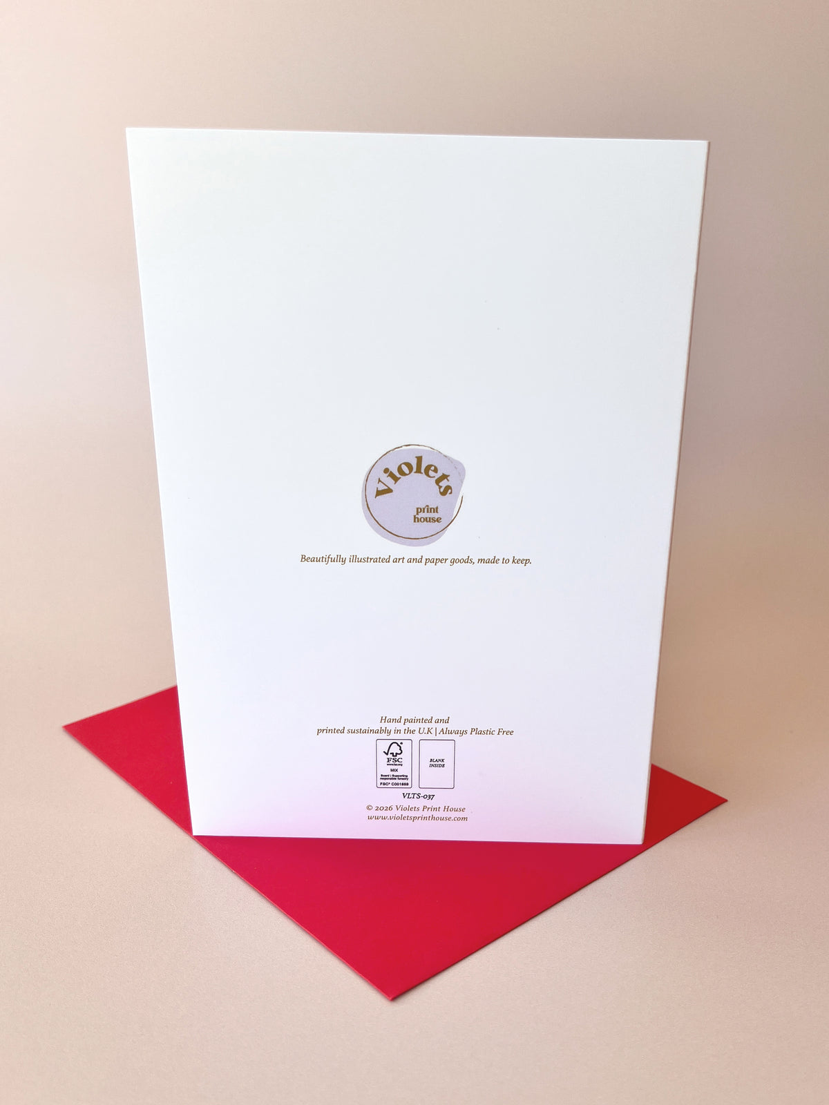 A white greeting card with a circular purple and gold 'Violeta' logo, standing upright on a red envelope against a warm beige background.