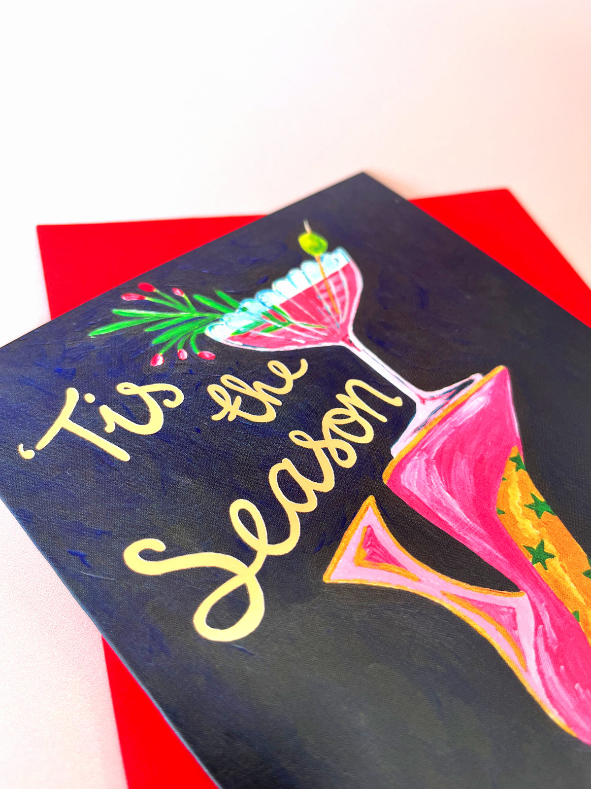 A square greeting card with a dark navy blue background featuring hand-painted illustrations of two colorful martini glasses and the text 'Tis the Season' in gold script. The card is layered on top of a bright red envelope against a clean white background.