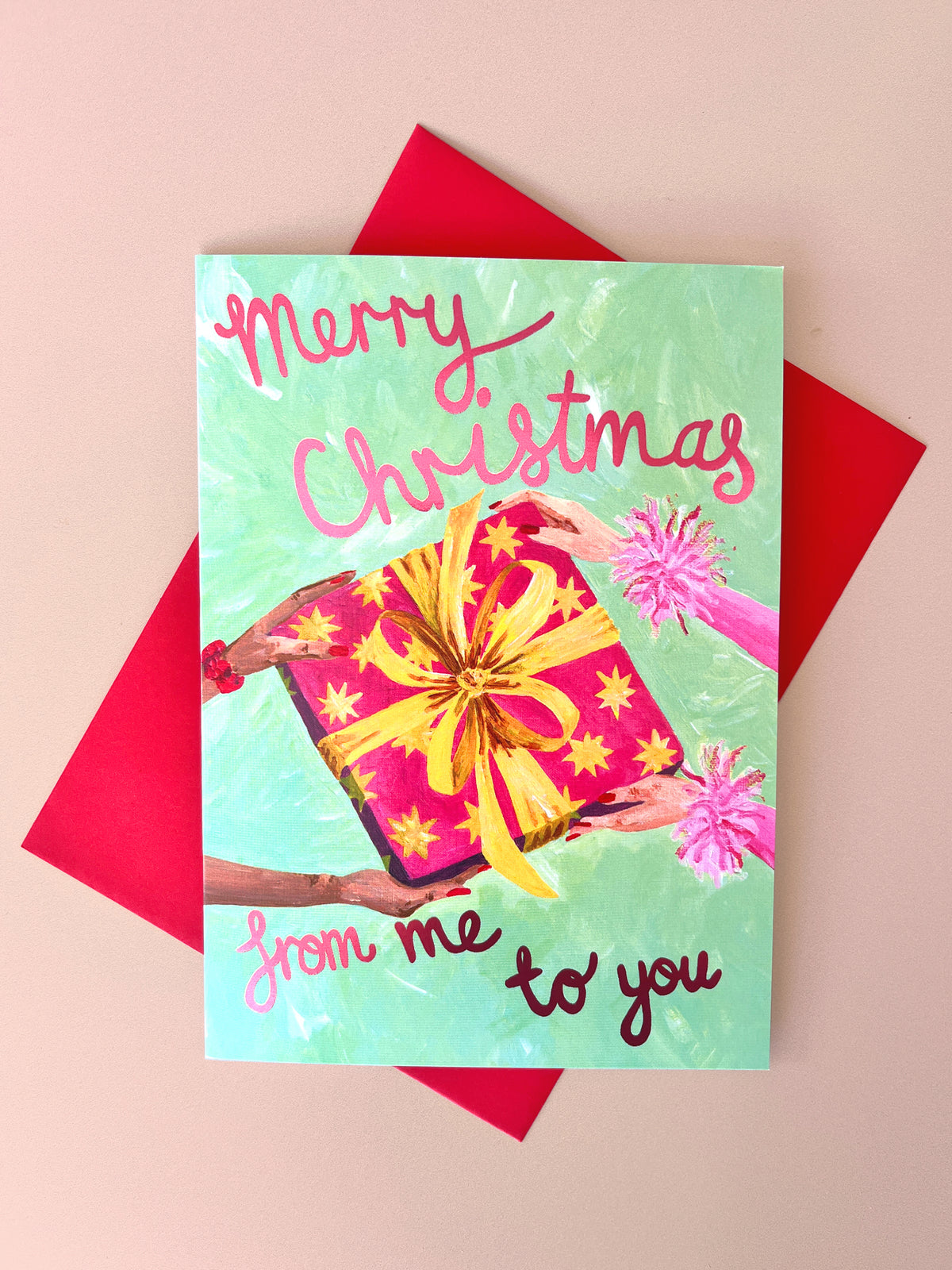 A square Christmas greeting card with a mint green background, featuring the text 'Merry Christmas from me to you' in pink and dark red script. The card is illustrated with hands holding a pink gift box with a yellow bow and pom-poms, and it is displayed on top of a red envelope against a light pink background.