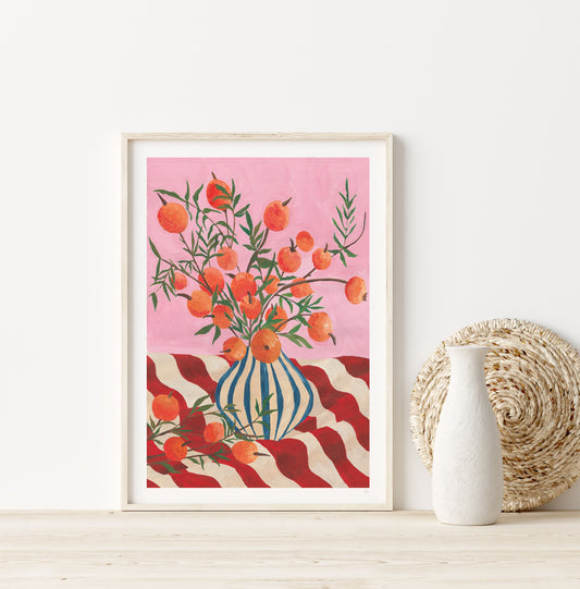 Framed artwork with a vase of oranges on a pink background, placed on a wooden surface.