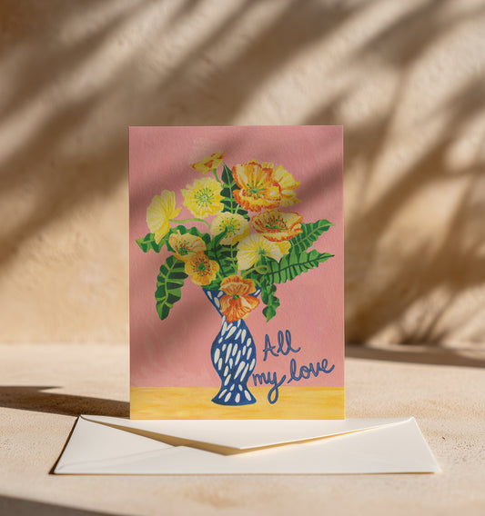 A greeting card with a floral illustration in a blue vase on a pink background, with the words 'all my love' in script, displayed upright with an envelope on a beige surface under warm leaf shadows.