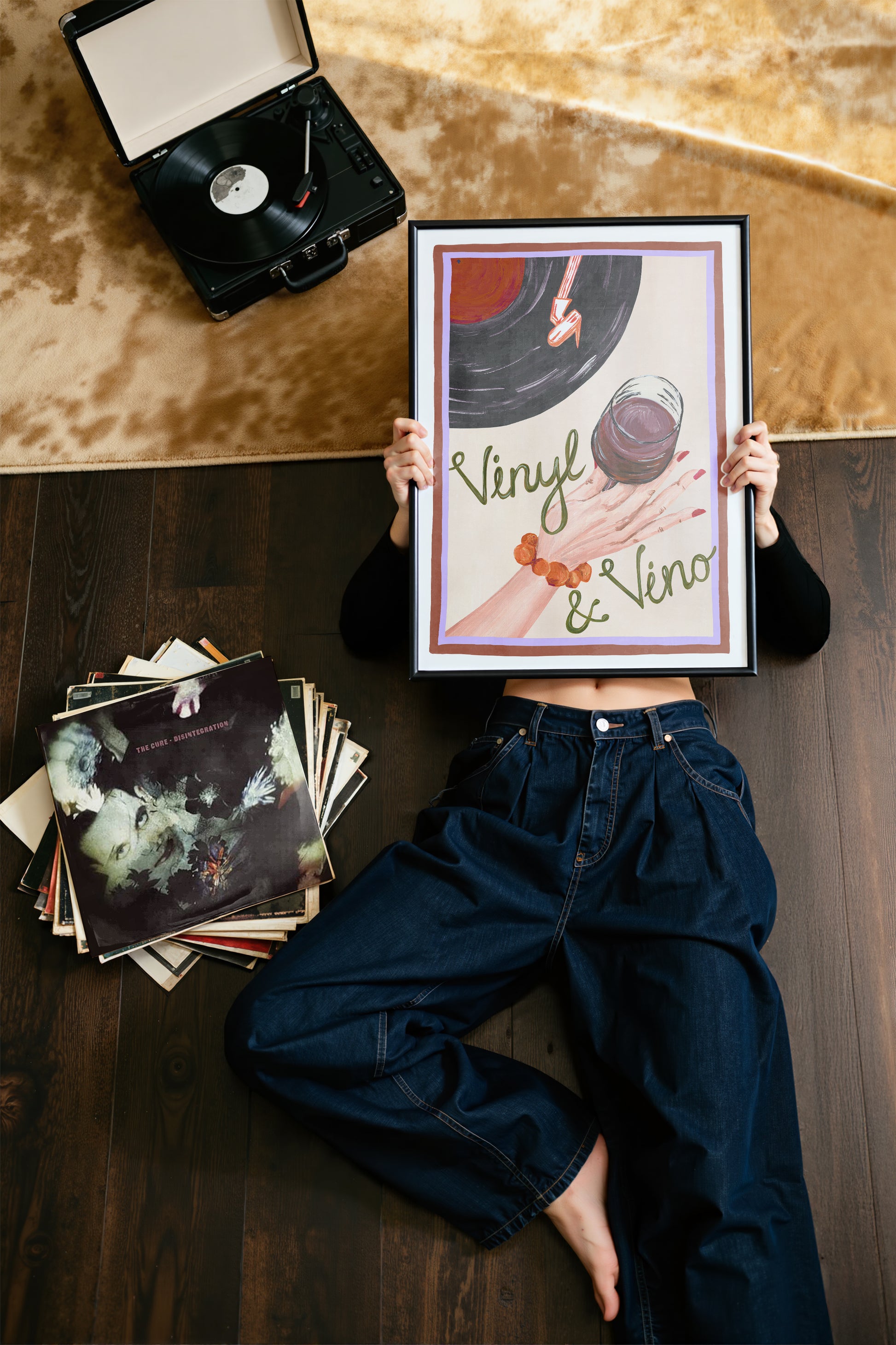 Person holding a framed vinyl record art print with records and a record player on a wooden floor.
