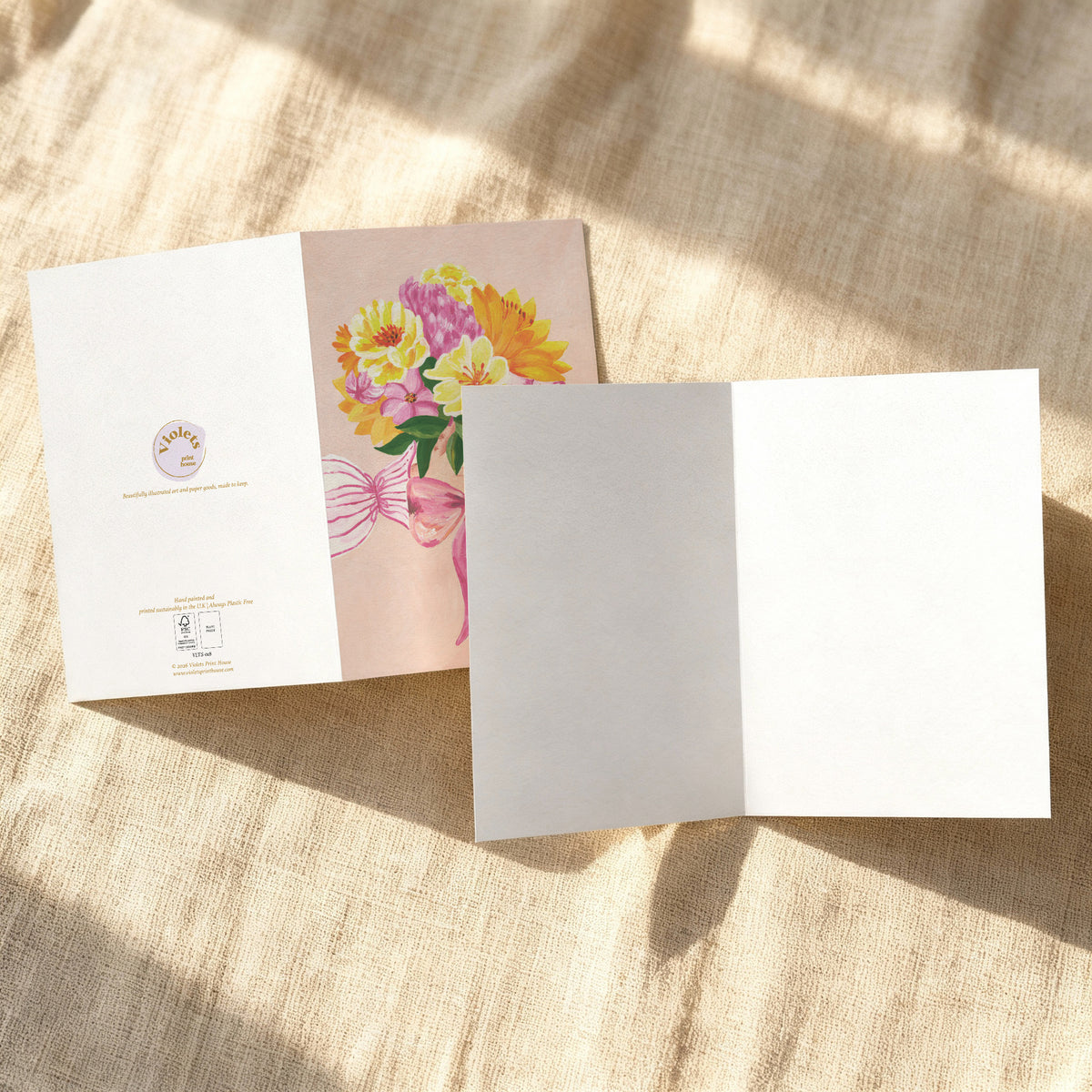 A folded white greeting card or invitation is shown on a textured beige fabric surface with soft sunlight and shadows. The card is partially open, revealing a floral illustration with yellow, pink, and orange flowers on a pale blush background.