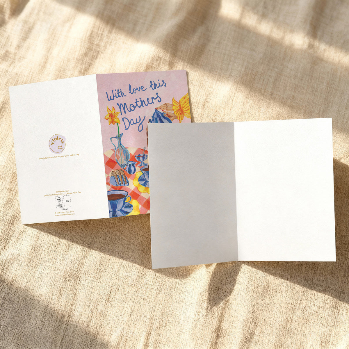 An open Mother's Day greeting card laid on a beige linen surface with dappled sunlight. The left panel shows a pastel illustration with the text 'With love this Mother's Day' and colorful floral and cup motifs.