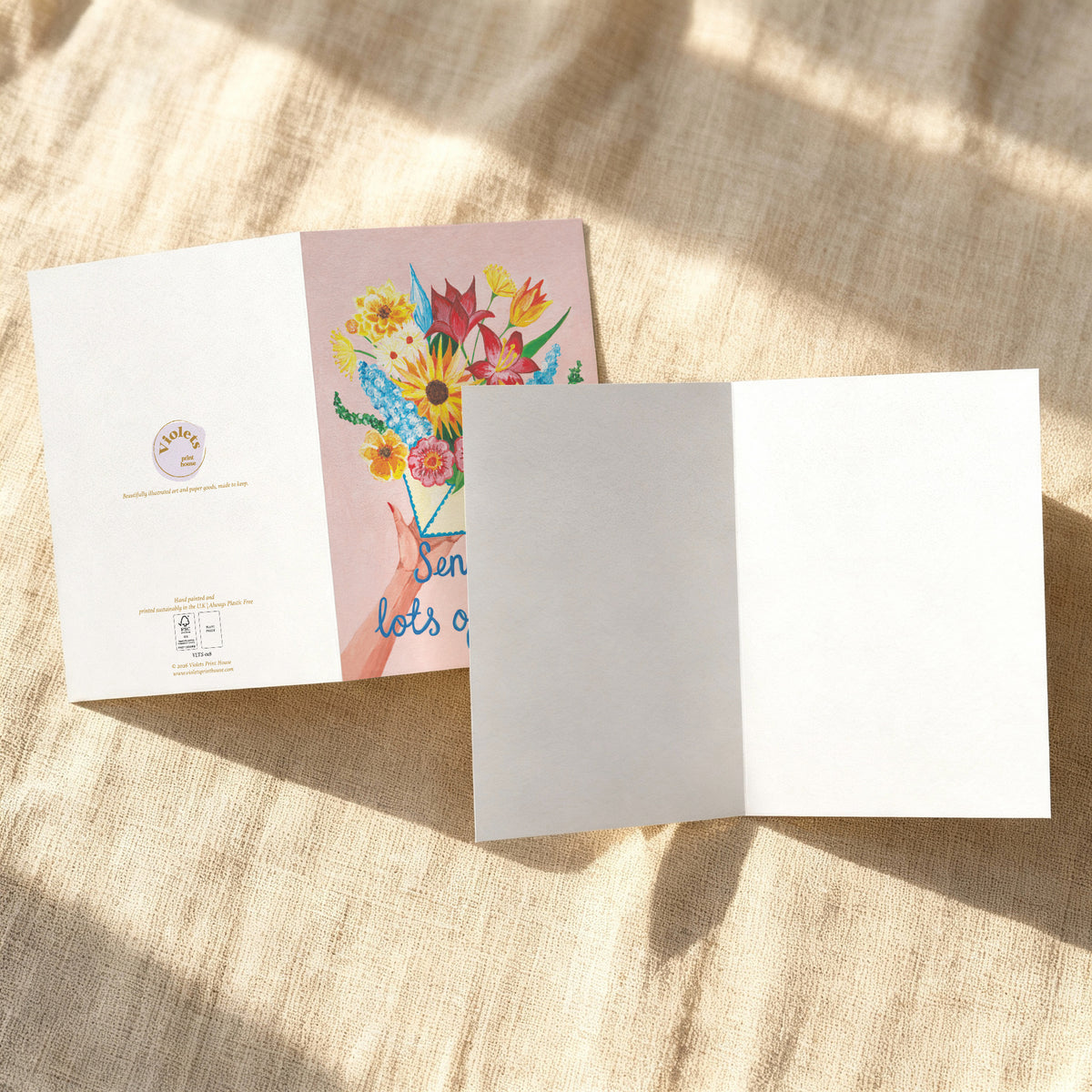 A folded greeting card shown open and closed on a beige linen surface with dappled sunlight. The left card displays a pastel pink floral illustration with colorful flowers and partial text, while the right card is plain white.