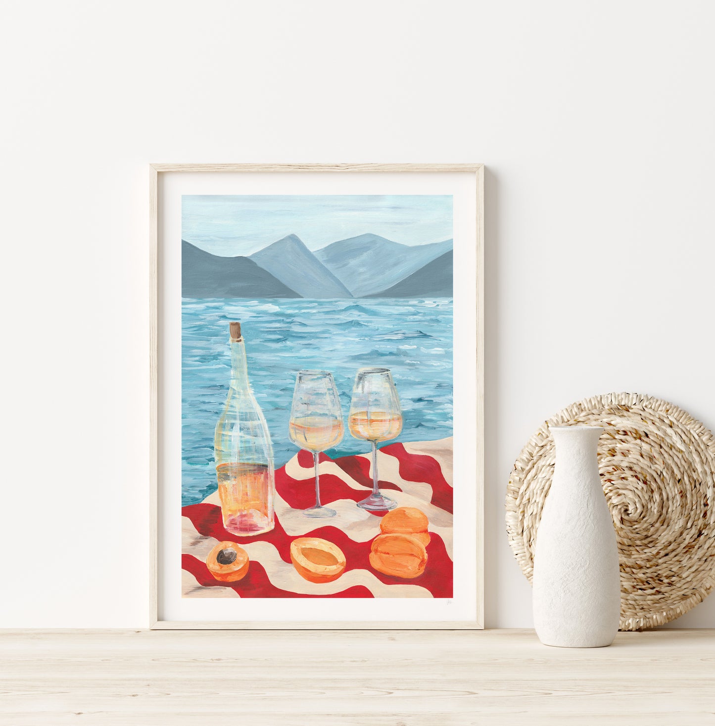 Framed artwork of a scenic beach scene with wine and macarons on a red blanket.