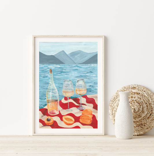 Framed artwork of a scenic beach scene with wine and macarons on a red blanket.