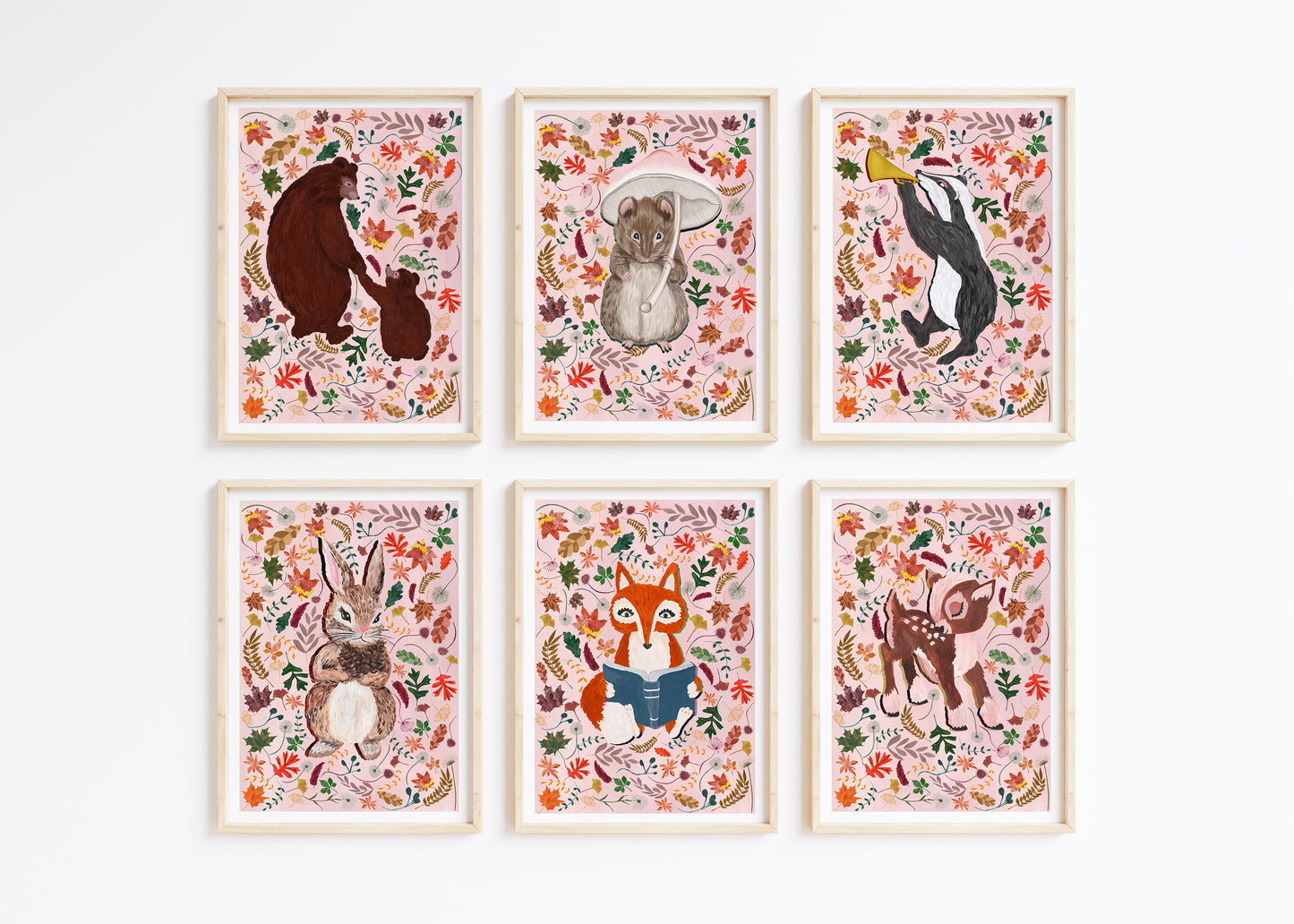 A set of 6 woodland animal prints, of a bear, mouse, badger, rabbit, fox and deer suurounds by autumn leaves on a pale pink background, framed on a white wall