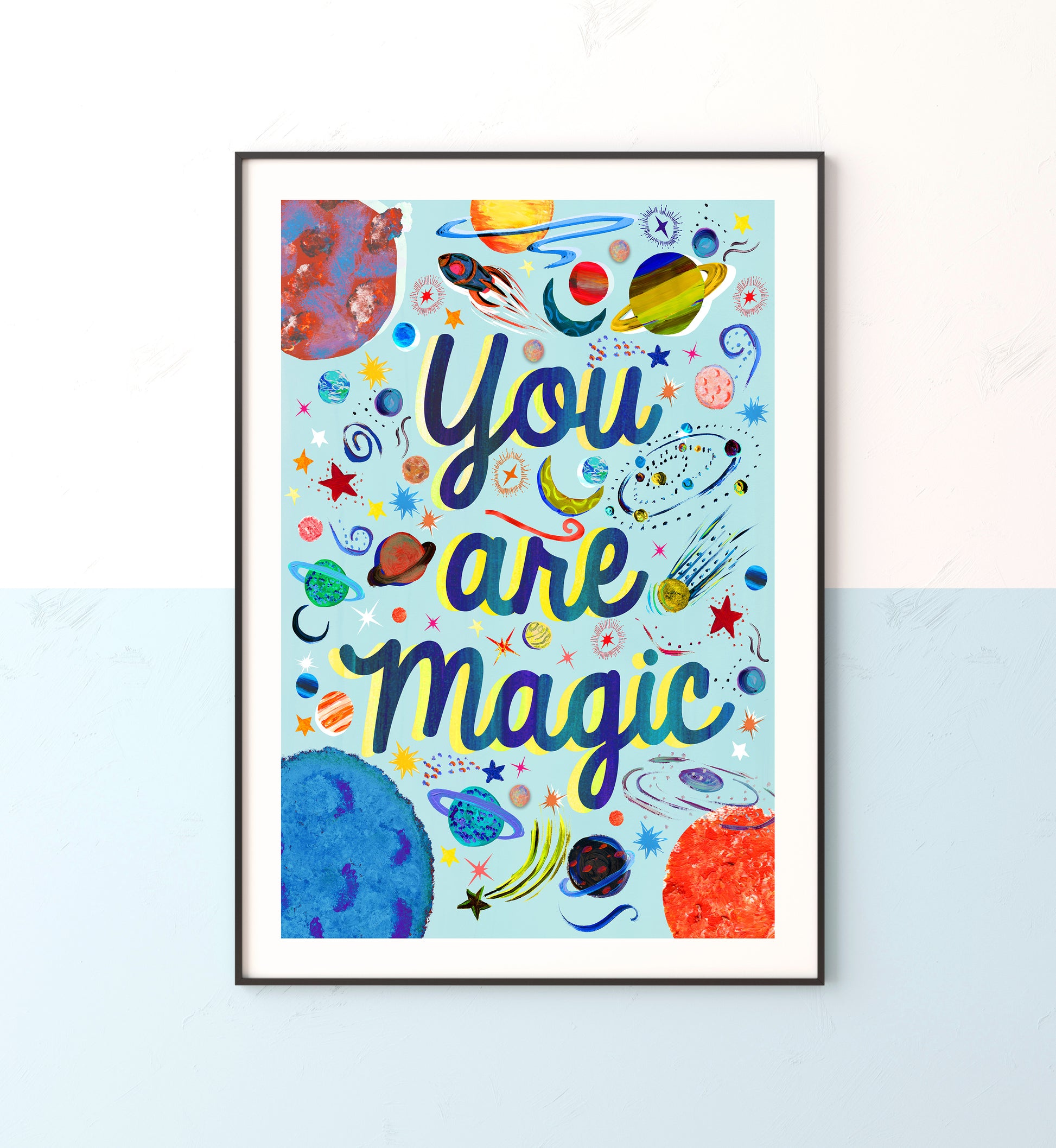 Space illustrated prints with planets and stars with teh words you are magic on a light blue background in a black frame on a blue and white wall