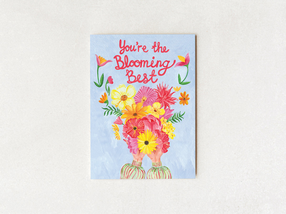 A square greeting card with a light blue background and the phrase 'You're the Blooming Best' in pink script, decorated with a colorful bouquet of flowers and illustrated hands at the bottom, shown against a light textured wall.