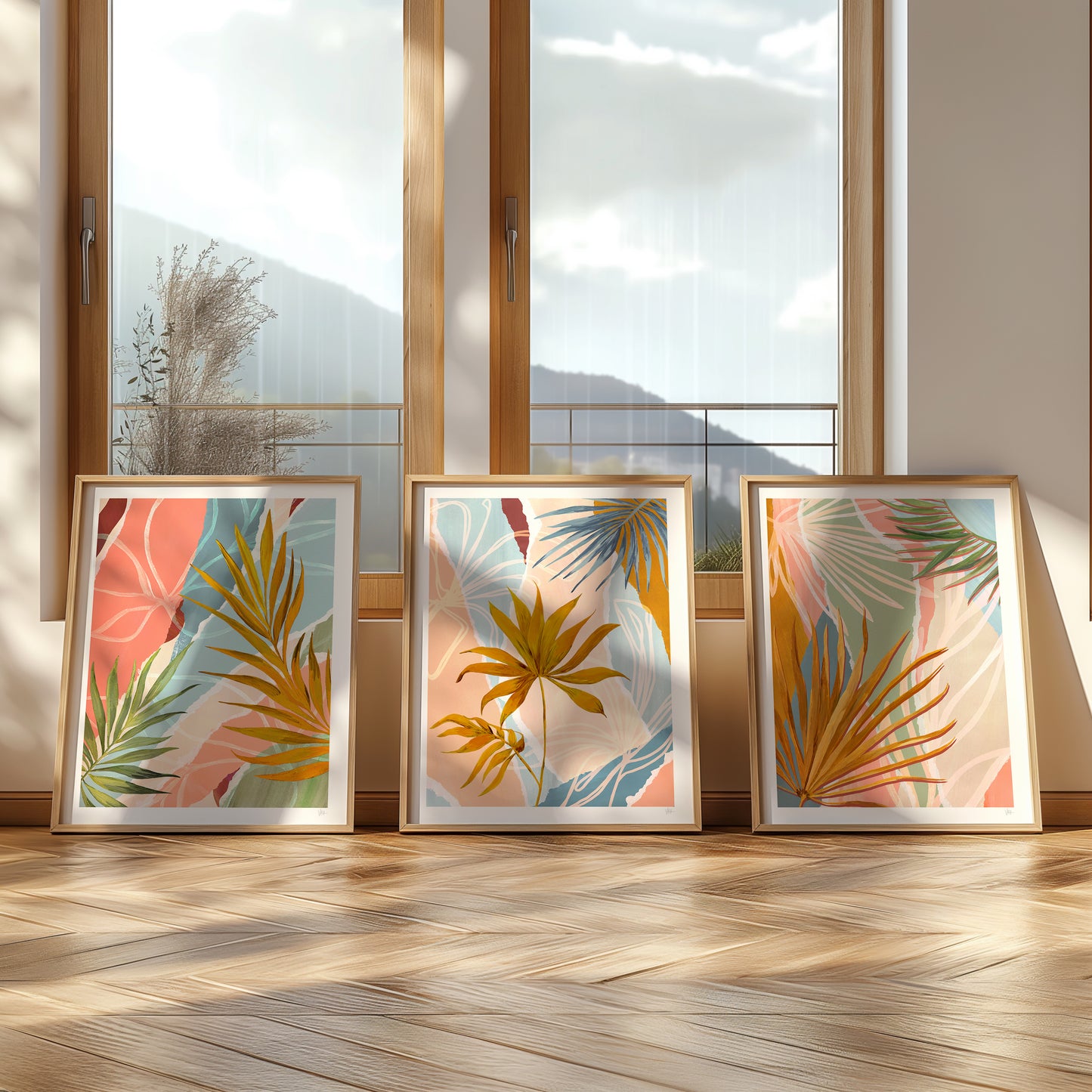 Three framed tropical leaf prints on a wooden floor with large windows in the background.