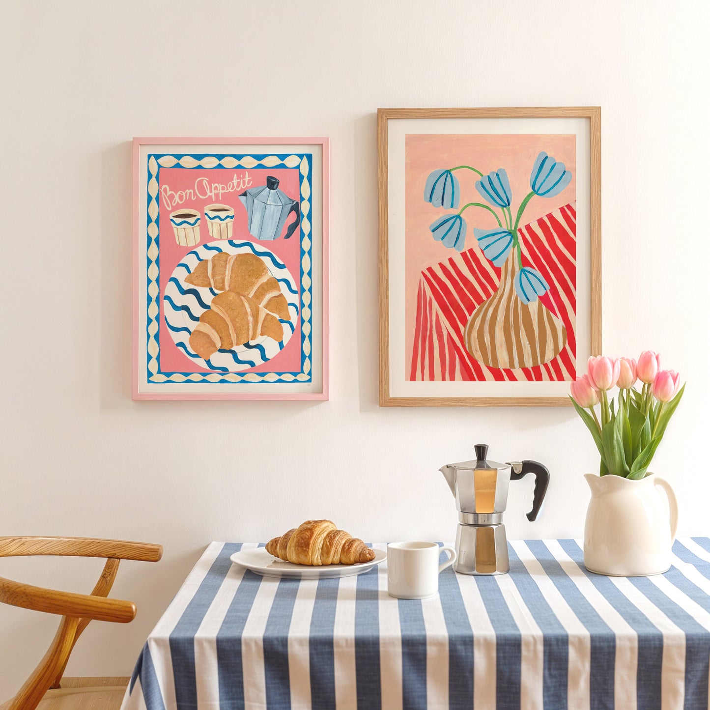 Two framed artworks on a wall above a table with a striped tablecloth, coffee maker, and flowers.
