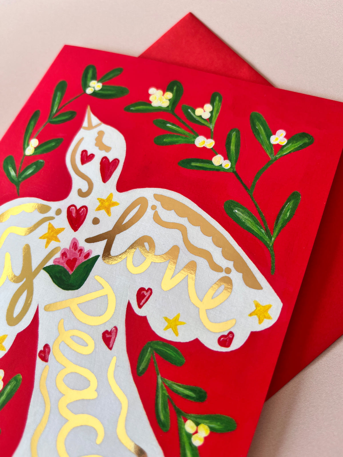 A close-up of a red greeting card featuring a white angel silhouette with gold foil script reading 'love' and 'peace', decorated with green leaves, small yellow flowers, and red hearts, with a matching red envelope behind it.