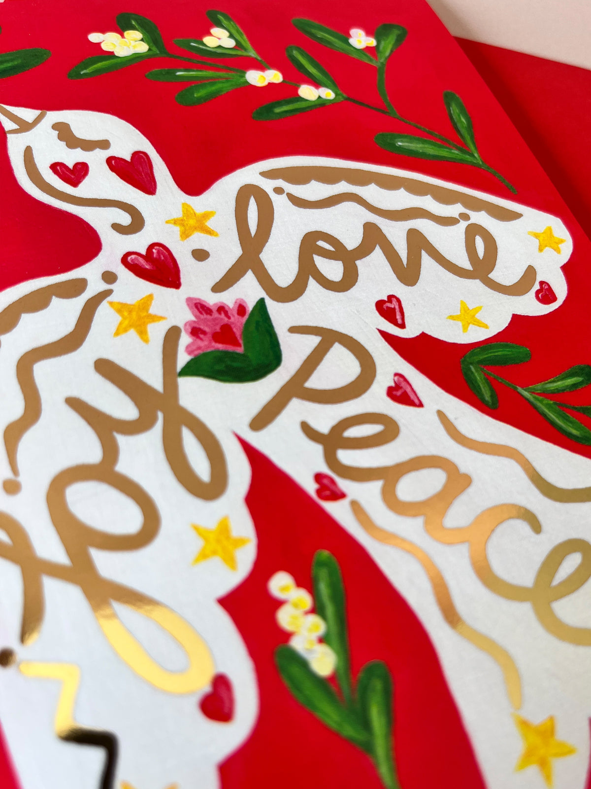 A close-up of a red greeting card featuring a white silhouette filled with the words “love peace joy” in gold script, decorated with small red hearts, yellow stars, and green floral branches.