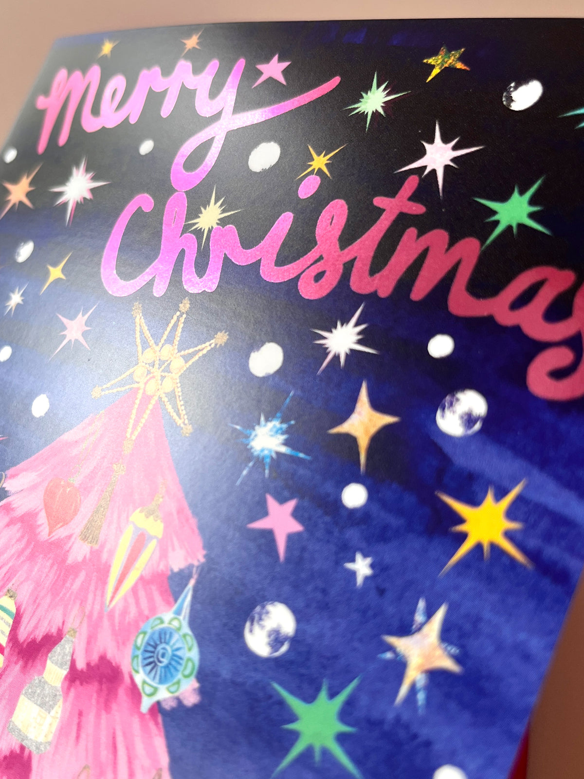 A close-up of a Christmas card featuring the words 'Merry Christmas' in pink script over a dark blue background decorated with colorful holographic stars and a pink tree illustration.