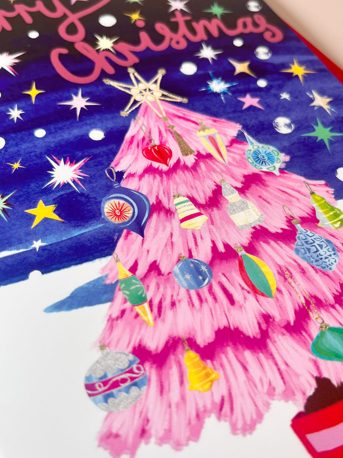 A close-up of a colorful Christmas card or print featuring a bright pink tree decorated with ornaments, a gold star topper, a blue watercolor sky with white and yellow stars, and pink script text reading 'Merry Christmas'.