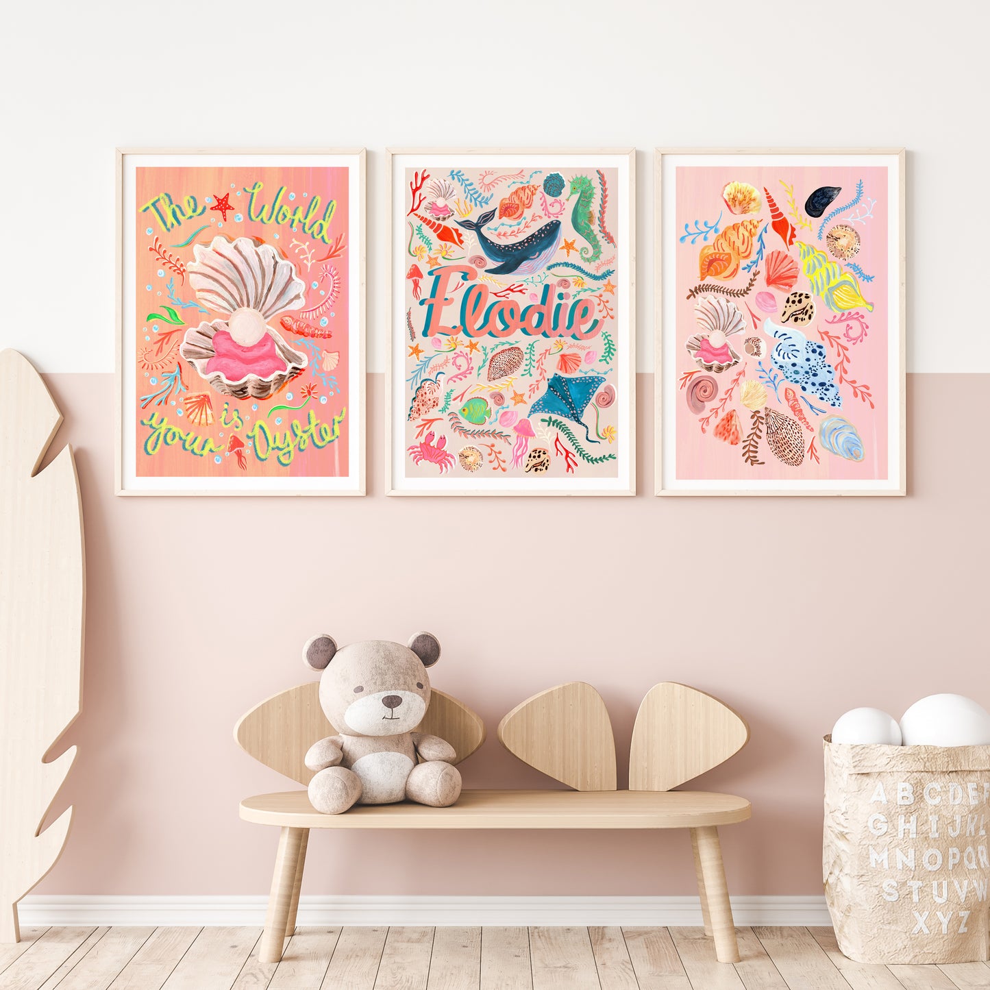 Three colorful framed ocean artworks on a wall one of 'the world is youy oyster' with a shell illustration, a name artwork with sealife illustrations and a selection of different shells illustration with a teddy bear and wooden bench below.