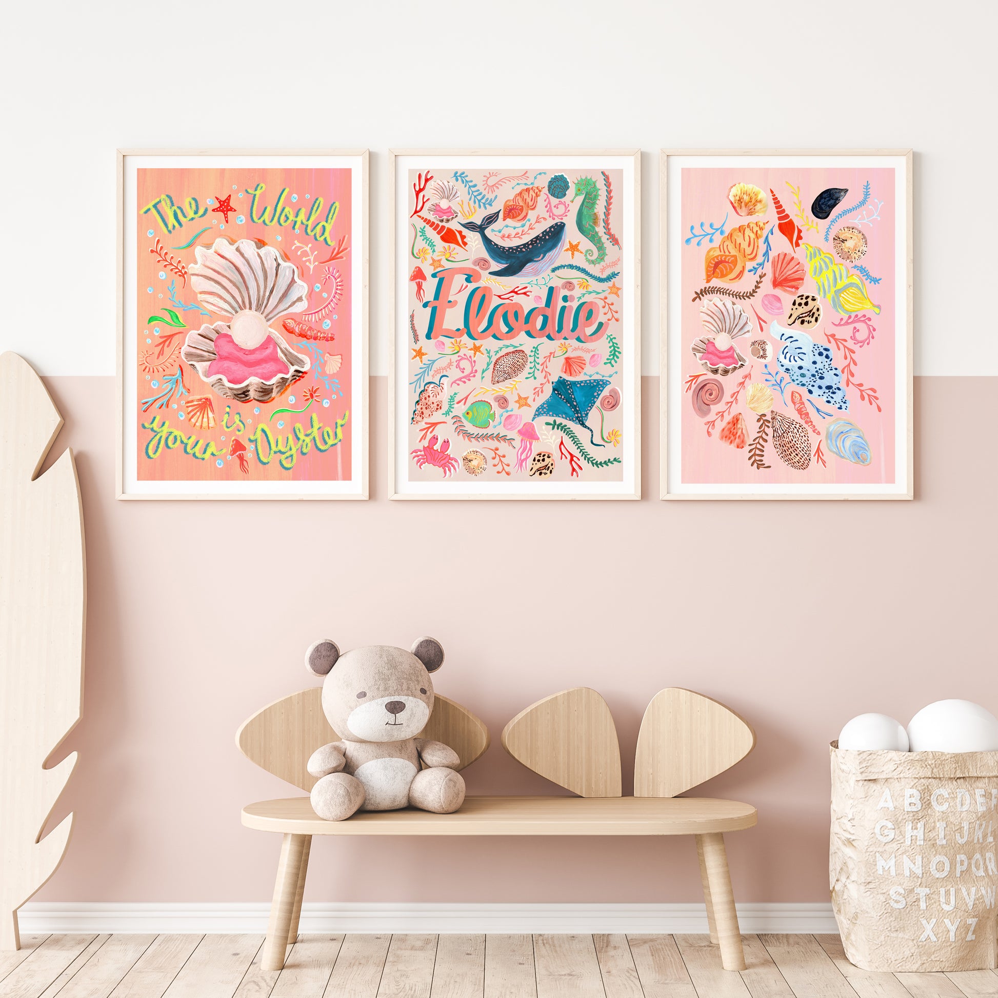 Three colorful framed ocean artworks on a wall one of 'the world is youy oyster' with a shell illustration, a name artwork with sealife illustrations and a selection of different shells illustration with a teddy bear and wooden bench below.