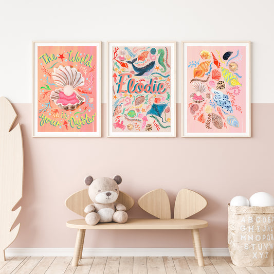 Three colorful framed ocean artworks on a wall one of 'the world is youy oyster' with a shell illustration, a name artwork with sealife illustrations and a selection of different shells illustration with a teddy bear and wooden bench below.
