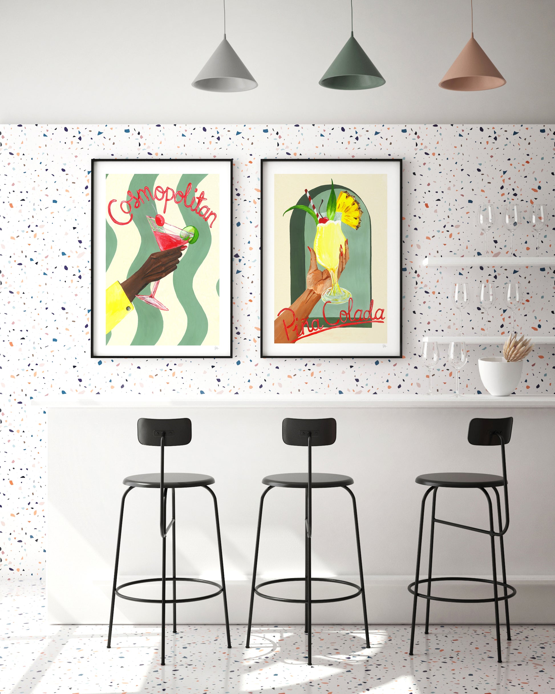 Two framed cocktail posters on a wall above three black bar stools in a room with a speckled white and gray wall.