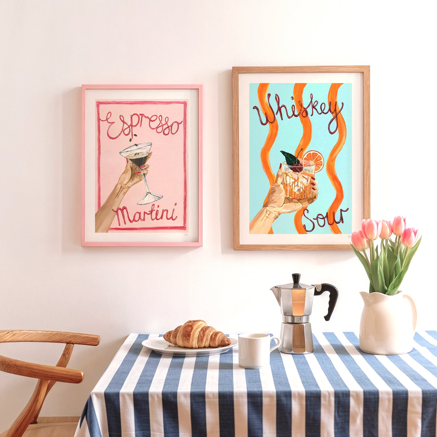 Two framed cocktail prints on a wall above a table with a coffee maker, croissant, and tulips.