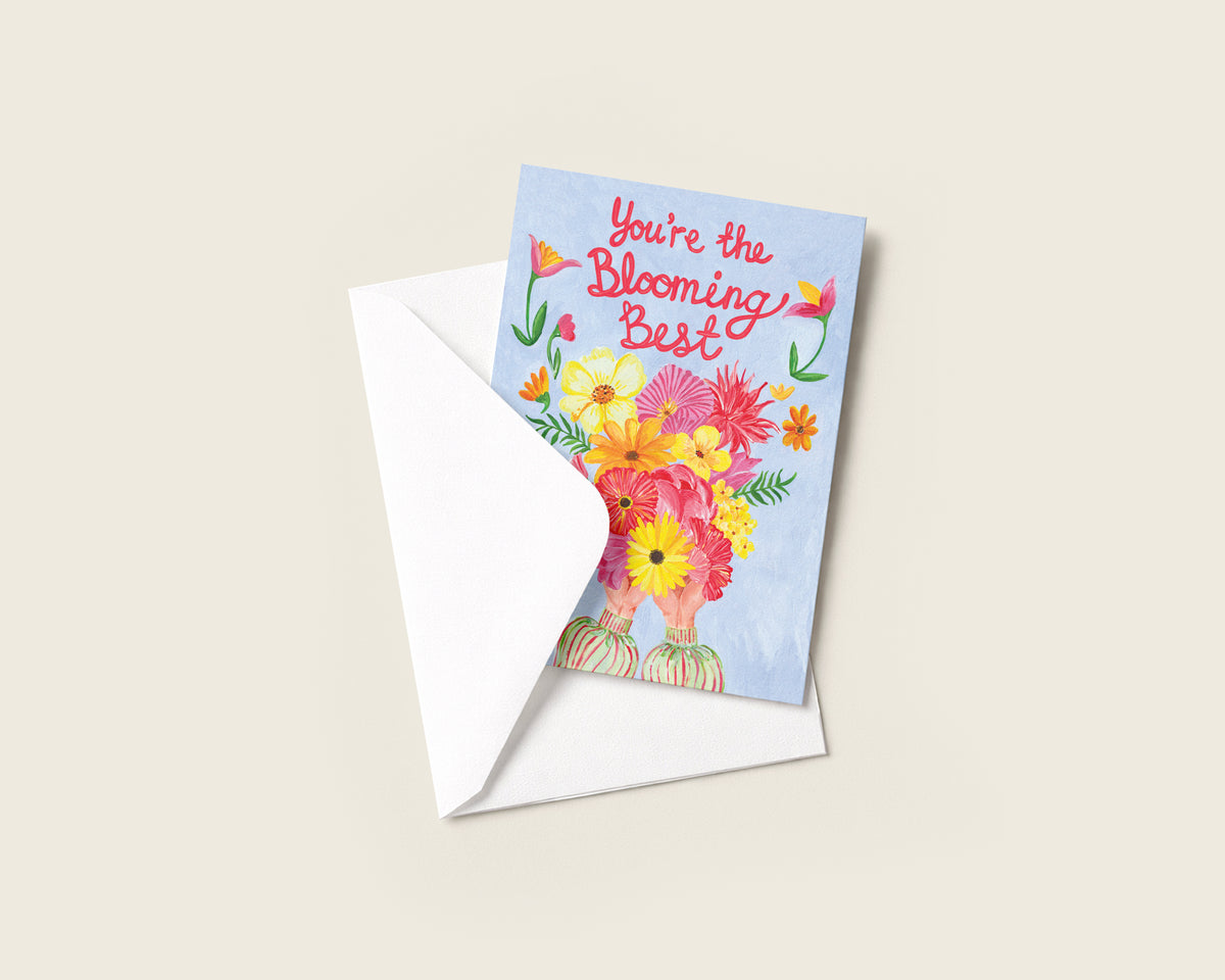 A square greeting card with a watercolor floral bouquet and the phrase 'You're the Blooming Best' in red script, shown with a white envelope on a light beige background.