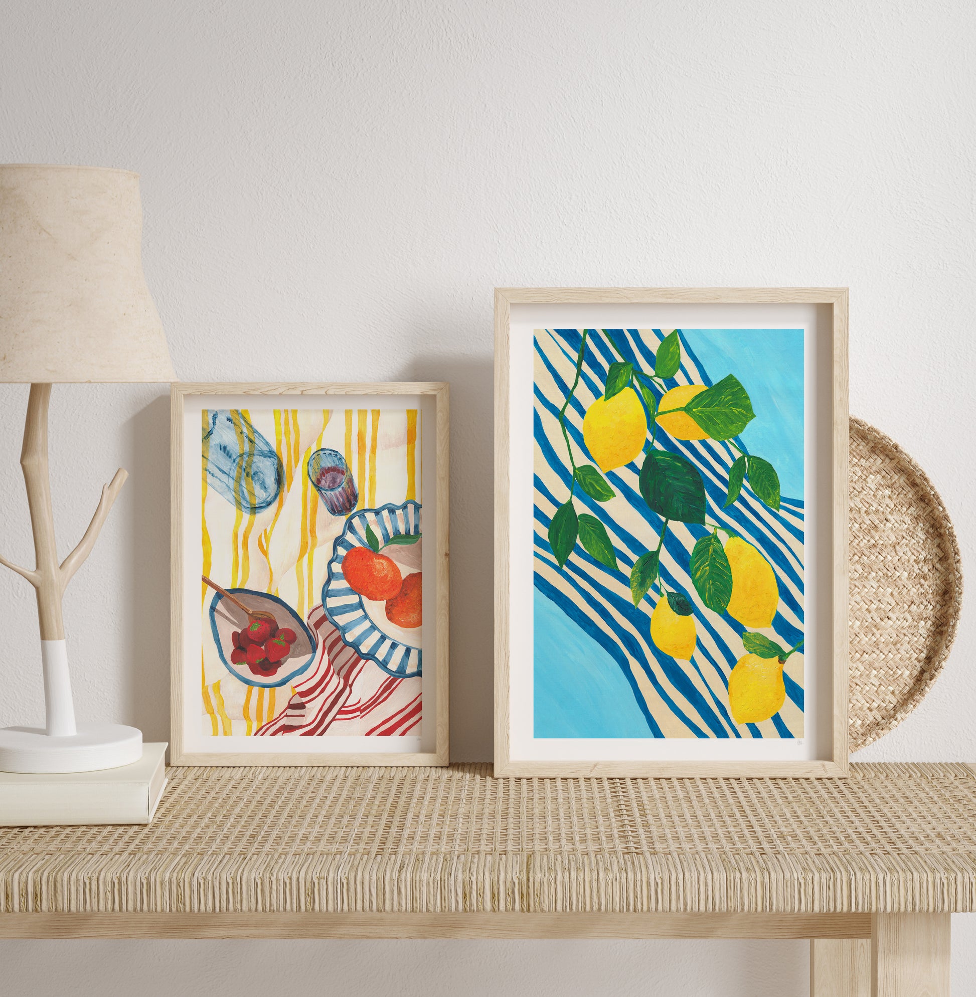 Two framed fruit on stripes artworks on a shelf with a lamp and decorative items.