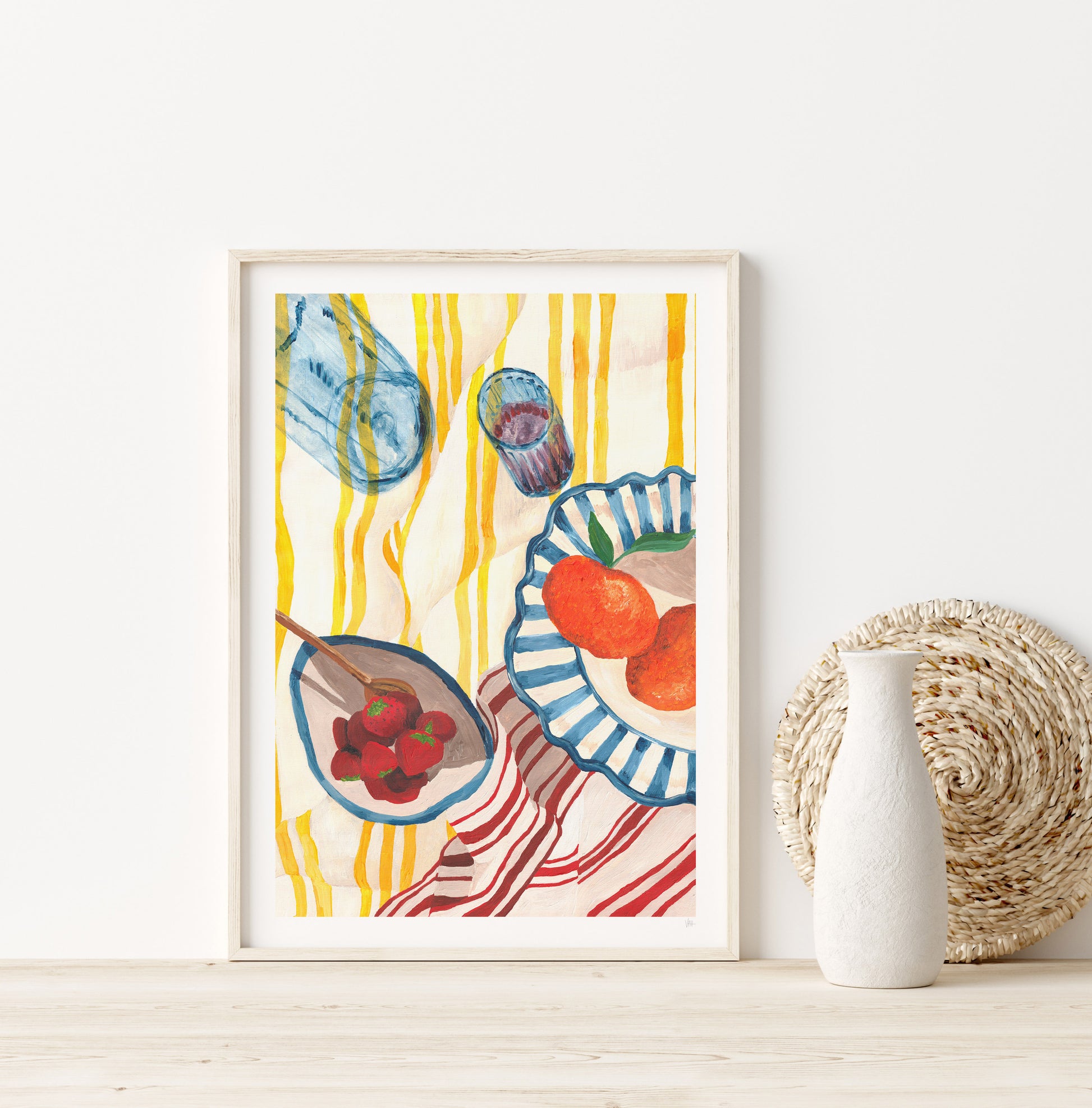 Framed colorful artwork of kitchen items on a white wall with a vase and woven basket.