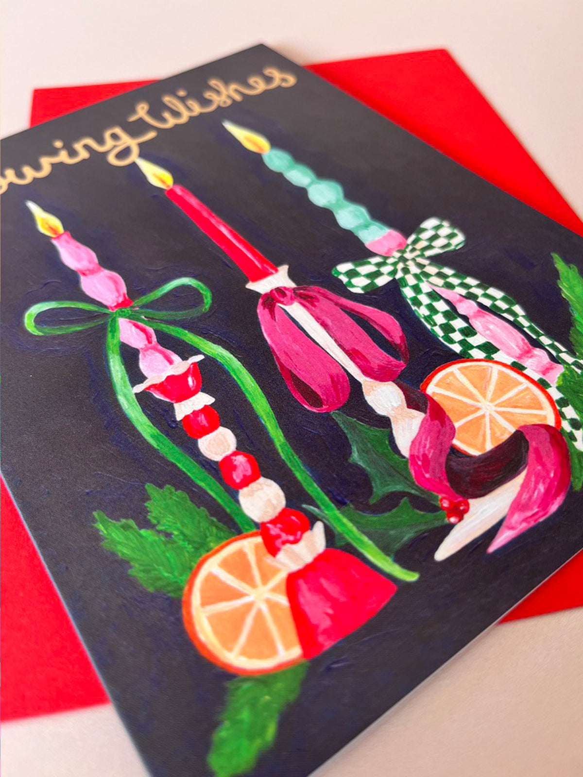 A square greeting card with a dark navy background and gold script reading 'Merry Wishes,' decorated with colorful painted candles, green leaves, red ribbon, and orange slices, placed on top of a red envelope.