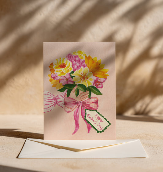 A Teacher's Day greeting card featuring a colorful watercolor bouquet of yellow, pink, and white flowers with green leaves and a pink ribbon, complete with a small 'Happy Teacher's Day' tag. The card is displayed upright on a cream-colored envelope on a sandy surface with warm natural light and leaf shadows.
