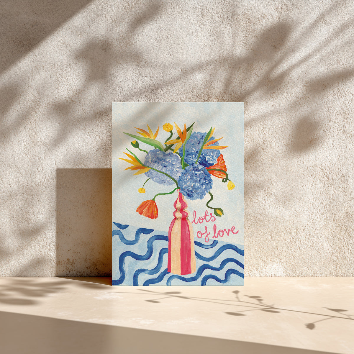 A vertical art print featuring a watercolor-style bouquet of blue, orange, and yellow flowers in a pink vase on a light blue background, with the words 'lots of love' in pink script. The print is displayed leaning against a textured beige wall on a sunlit surface with leaf shadows cast across it.