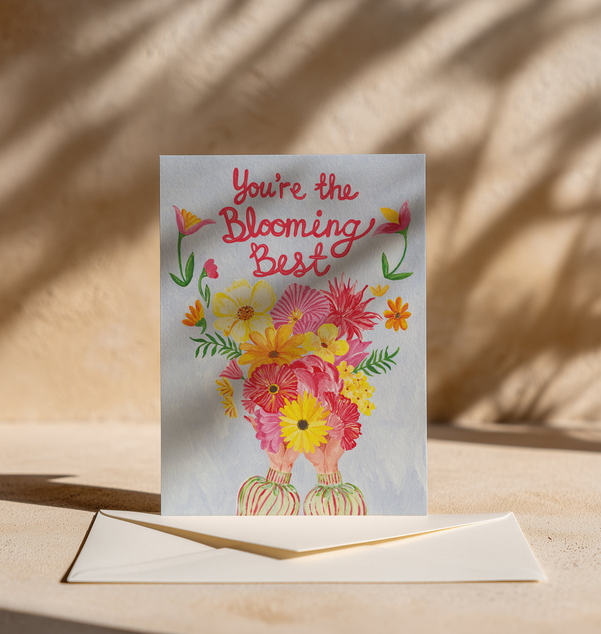 A greeting card with the phrase 'You're the Blooming Best' in red script above a colorful bouquet illustration, displayed upright on a beige surface with an envelope in front and warm shadow patterns in the background.