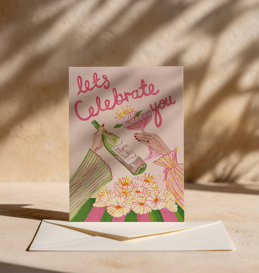 A greeting card with an illustration of hands holding a wine bottle and a glass of red wine, surrounded by flowers, with the text 'let's Celebrate you' in pink script. The card is displayed upright on a cream envelope on a beige surface with warm, dappled sunlight and shadows.