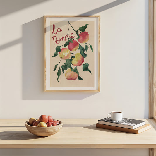 Framed artwork of apples with 'la Pomme' text on a wall above a wooden table with a bowl of apples and a cup.