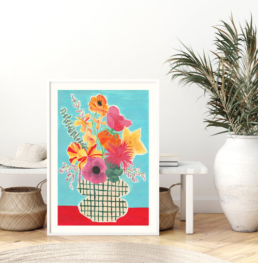 Colorful floral artwork in a white frame on a wall with a plant and decor items below.