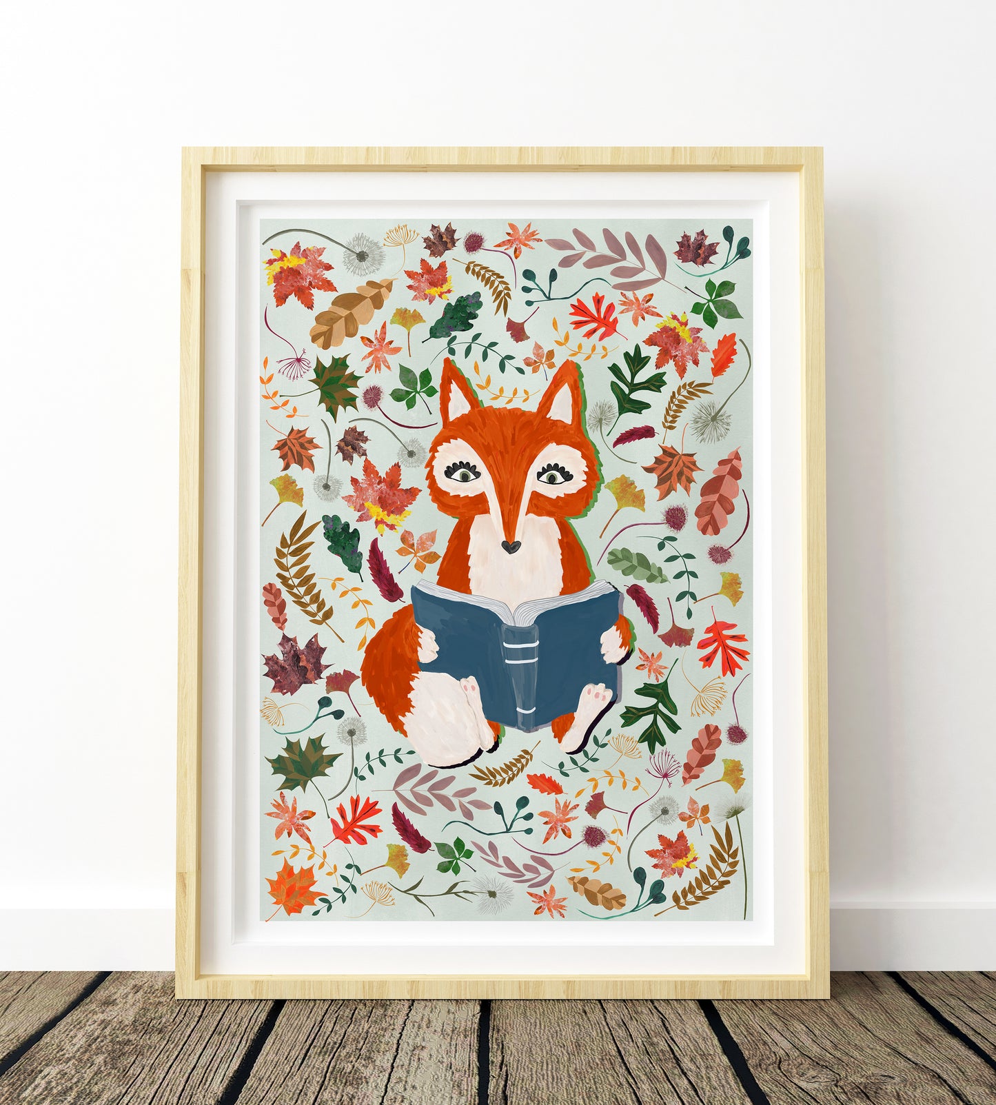 Framed artwork of a fox reading a book surrounded by autumn leaves on a white wall.