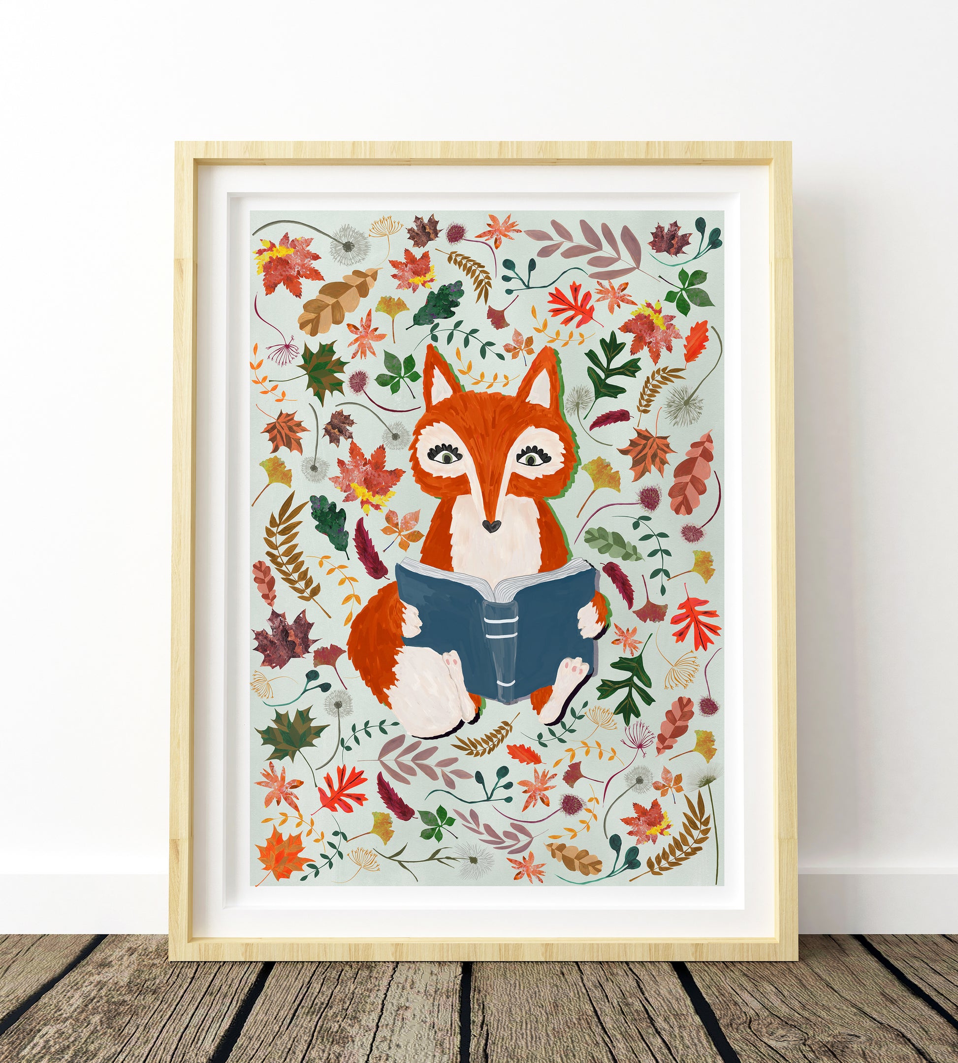 Framed artwork of a fox reading a book surrounded by autumn leaves on a white wall.