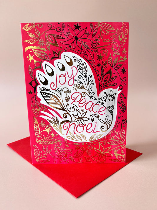 A red greeting card with a white hamsa hand and gold foil details, reading 'Joy Peace Noel' in pink script, displayed with a matching red envelope on a beige background.