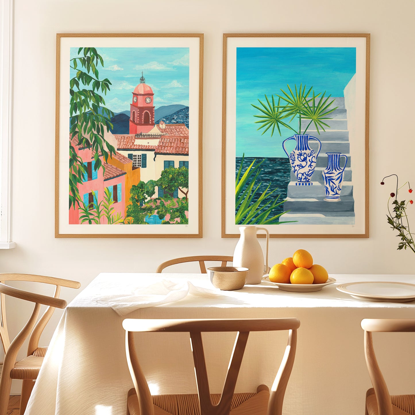Two framed artworks on a wall above a dining table with fruit and a pitcher.