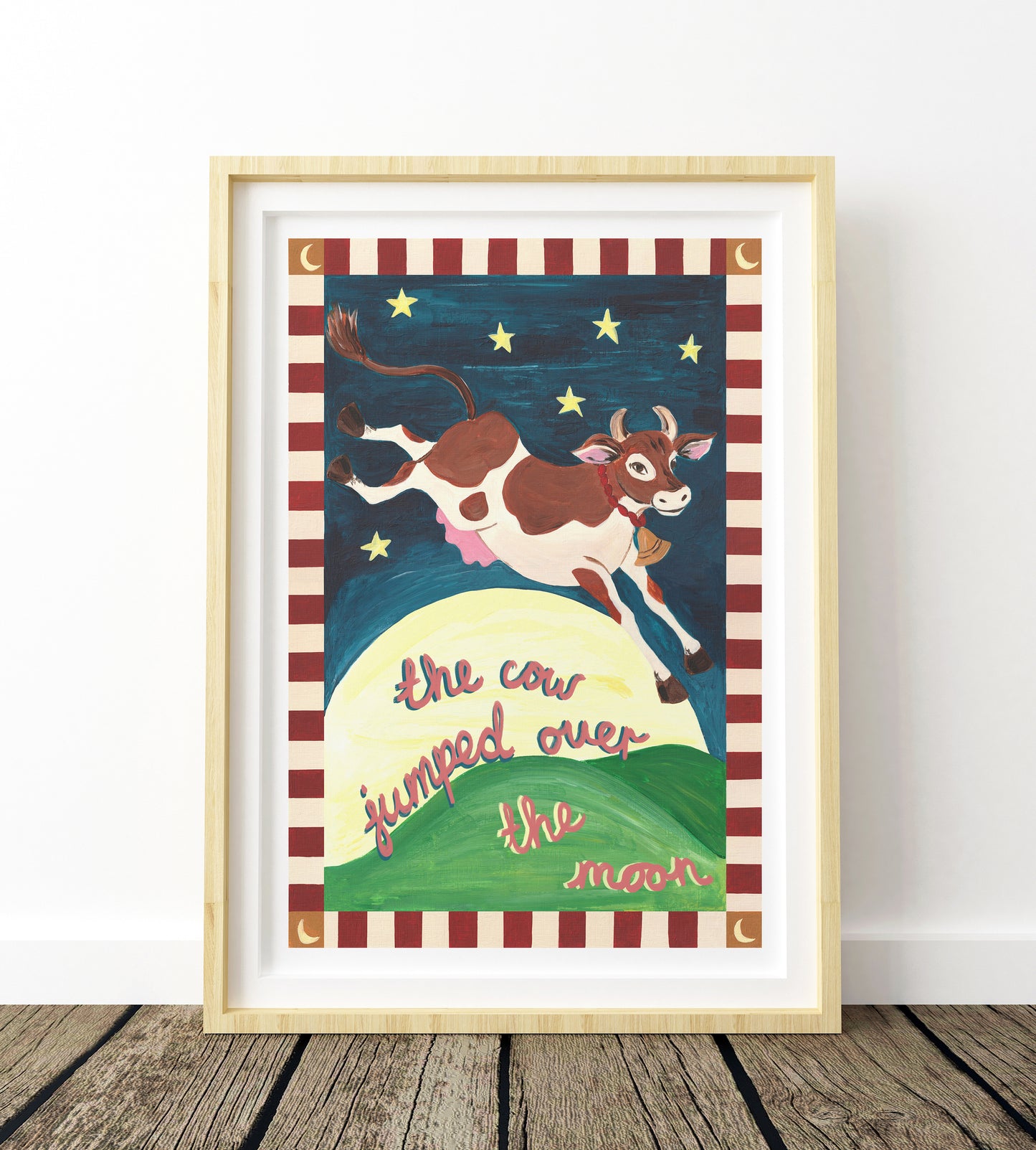 Framed artwork of a cow jumping over the moon with a striped border on a wooden floor.