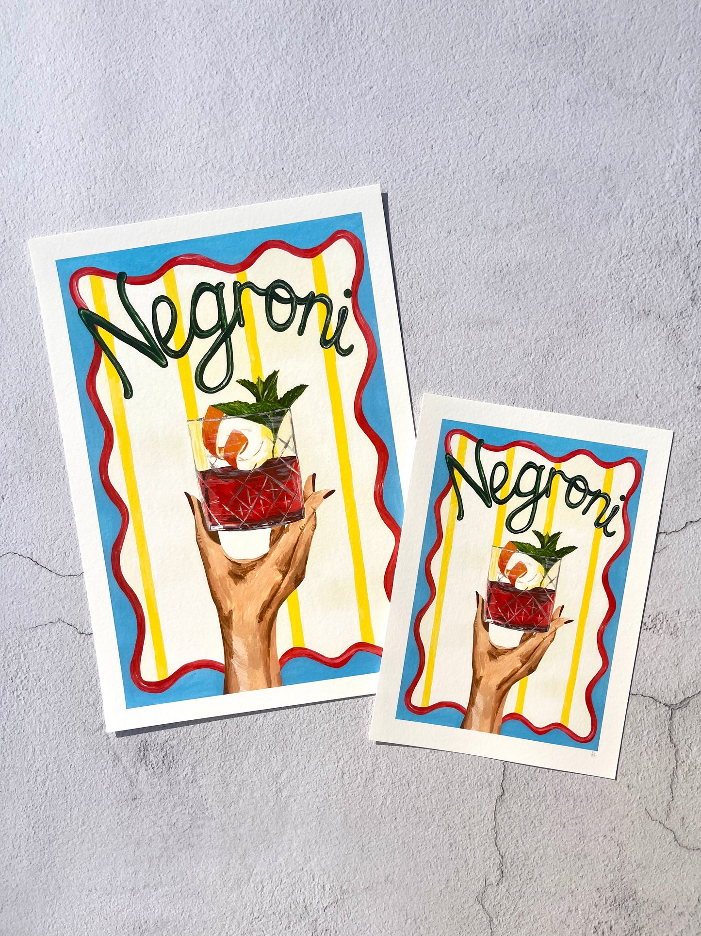 Two Negroni-themed prints with a hand holding a glass of Negroni on a gray background.